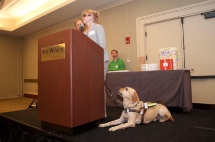 Jen Buchanan speaking at a podium at a MABVI Volunteer Appreciation Brunch, with her guide dog Keating on the ground looking up at her