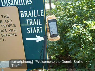 A photo of a smartphone in the LaunchGuide location, with a sign reading "BRAILLE TRAIL START" pointing to the phone in the wire guides and a caption on the photo that reads "[smartphone:] Welcome to the Dennis Braille Trail"