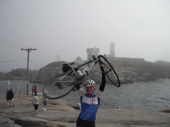 Megan in biking gear holding her bicycle above her head by the ocean with a lighthouse in the background